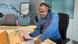 A DW reporter wearing headphones sits at a desk in a DW radio studio in Bonn, Germany, speaking into a microphone and reading from a sheet of paper.