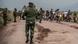 Motorcycles carrying people and their belongings on a road patrolled by soldiers